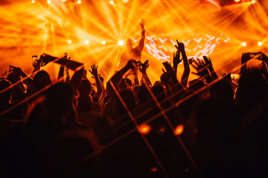 Silhouettes of people rising their hands and dancing on on open air music festival at night while performer on stage is making a good atmosphere. There are orange stage lights. Nightlife, concerts.